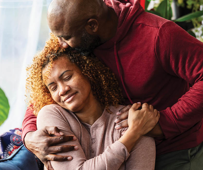 A mature man shows his wife how much he love her, wrapping his arms around her shoulders to give her a hug, and kissing her on the side of her head. She is smiling with her eyes closed, resting her hand on his.